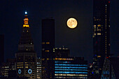 View of a radiant full moon glows ethereally behind the illuminated cityscape, casting long shadows across modern skyscrapers, New York, New York, United States.