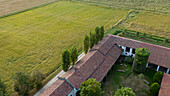 Aerial view of the rustic farmhouse, its terracotta roof glowing warmly against the lush green fields, a serene pastoral scene unfolding from above, Vespolate, Piedmont, Italy.