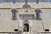 View of the imposing stone fortress entrance, adorned with flags, intricate carvings and a drawbridge, under the bright sun, L'Aquila, Abruzzo, Italy.