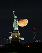 View of The Statue of Liberty standing tall against the dark night sky, with a reddish moon looming behind, a serene yet powerful scene, New York, New York, United States.