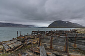 View of weathered, decaying wooden wreckage scattered along the shore, with the serene turquoise waters meeting distant, mist-shrouded mountains, Longyearbyen, Svalbard, Svalbard and Jan Mayen.