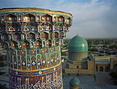 Aerial view of vibrant blue and white mosaic patterns adorn the towering structure, contrasting with the soft green dome in the distance, Samarkand, Uzbekistan.
