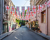 View of Georgian flags flutter above a narrow street lined with weathered buildings and vibrant graffiti, casting shadows on the asphalt, Tbilisi, Tbilisi, Georgia.