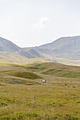 Blick auf goldene Wiesen, die sich unter einem blassen Himmel auf ferne, dunstige Berge zubewegen, mit einer kleinen Struktur im Tal, Isola del Gran Sasso D'italia, Abruzzen, Italien.
