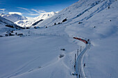 Aerial view of a vibrant red train slicing through the pristine white snow-covered landscape near the majestic mountains, Filisur, Switzerland.