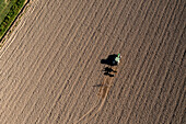 Aerial view of a lone tractor etching precise lines into the expansive, textured earth, creating a stark contrast against the green grass, Vespolate, Piedmont, Italy.