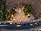 Aerial view of a cozy outdoor cafe nestled between lush trees, soft light spilling onto tables as cars blur past, Chania, Chania, Greece.