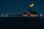 View of a radiant crescent moon hanging above a brick building, its lights mirroring on the dark waters in the tranquil night, New York, New York, United States.