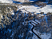 Aerial view of a winding road cutting through a snowy forest contrasting with open snowfields and distant buildings under a bright sun, Maloja Pass, Maloja, Switzerland.