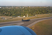 Aerial view of an airplane's shadow cast dramatically over the runway as it prepares for landing amidst a backdrop of verdant trees, Warsaw, Masovian Voivodeship, Poland.