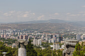 View of a sprawling cityscape nestled between verdant trees and distant mountains, framed by architectural stone column details, Chronicle of Georgia, Tbilisi, Georgia.