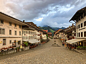View of cobblestone streets flanked by buildings with warm lights under a dramatic sky, casting long shadows in Gruyères, Fribourg, Switzerland.