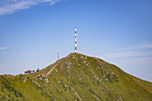 Blick auf einen markanten rot-weißen Turm, der den azurblauen Himmel auf einem grünen, zerklüfteten Berggipfel durchdringt, Goldeck, Kärnten, Österreich.