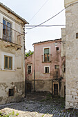 Blick auf verwitterte Gebäude mit verblassten rosafarbenen und cremefarbenen Fassaden, die eine Kopfsteinpflasterstraße unter einem klaren Himmel einrahmen, Bominaco, Abruzzen, Italien.