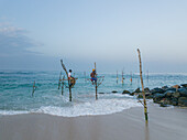 Aerial view of stilt fishermen perched on poles amidst the crashing waves, a timeless tradition against the serene ocean horizon, Weligama, Sri Lanka.