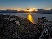 Aerial view of the sun reflecting off the water near the Palamidi fortress and the city, casting a golden glow over the historic landscape, Nafplion, Argolis, Greece.
