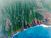 Luftaufnahme von zerklüfteten Klippen, die auf den türkisfarbenen Ozean treffen, ein Teppich aus Grün und Braun unter einem Nebelschleier, Napali, Hawaii, Vereinigte Staaten.