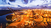 Oakland, United States - 11 May 2020: Aerial view of the Port of Oakland glows with warm light against the cool blues of twilight, the cranes and cargo ships standing as monuments to global trade.