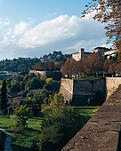 View of ancient stone walls embrace verdant hills where buildings nestle, bathed in warm sunlight under a sky brushed with clouds, Bergamo, Lombardy, Italy.