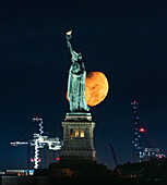 Blick auf die ikonische Freiheitsstatue, die sich vor einem riesigen, leuchtenden Mond abhebt, ein Leuchtfeuer der Freiheit am Nachthimmel, New York City, New York, Vereinigte Staaten.