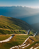 Aerial view of a winding road snaking through vibrant green hillsides touched with melting snow, contrasting against a distant, hazy mountain range, Sosan, Himachal Pradesh, India.