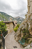 View of a narrow cobblestone path winds past rustic stone buildings clinging to a rocky hillside under a cloudy sky, Castrovalva, Abruzzo, Italy.
