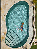 Aerial view of a lone figure floats serenely in a turquoise pool, surrounded by pale stone and verdant grass, Tambon Sam Roi Yot, Chang Wat Prachuap Khiri Khan, Thailand.