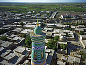 Aerial view of the vibrant turquoise and gold minaret rising above the ancient city's maze of flat-roofed buildings, a contrast to the distant green fields, Khiva, Uzbekistan.