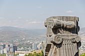 View of a weathered stone column fragment stands boldly in the foreground, overlooking the sprawling cityscape nestled among distant, hazy mountains, Chronicle of Georgia, Tbilisi, Georgia.