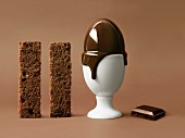 Zwei Streifen Schokoladenkuchen, schmelzendes Ei im Eierbecher und Schokoladenstück