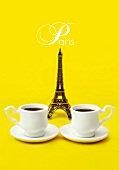 Komposition mit Mini-Eiffelturm und Kaffeetassen vor gelbem Hintergrund