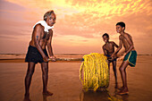 Fischer tragen ein aufgerolltes Seil bei Sonnenuntergang am Strand, Calangute Beach, Goa, Indien