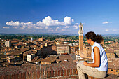 Woman overlooking city rooftops with Piazza del Campo and Torre del Mangia, Siena, Tuscany, Italy