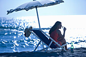 Woman sitting in deck chair on beach and drinking, Apulia, Italy
