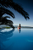 Woman sitting at edge of outdoor pool, sea in back, Bay of Porto Vecchio, Southern Corse, France