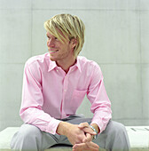 blonder Mann sitzt auf Steinbank, Deutscher, rosa Hemd, stylisch, modern, lächeln, freundlich, gelaltene Hände, sitzt vor heller Wand