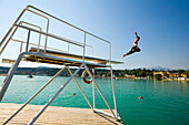 Boy jumping from diving platform into Worthersee, Hotel Schloss Velden in background, Velden, Carinthia, Austria