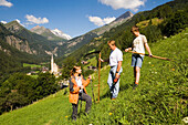 Woman talking with a man and a boy, man holding a scythe, Heiligenblut with pilgrimage church Zum hl. Pluet and Grossglockner in background, Heiligenblut, Carinthia, Austria