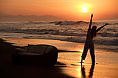 Eine junge Frau dehnt sich am Strand bei Sonnenuntergang, nahe Uluwatu, Bali, Indonesien