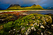 The midnight sun shines on the coastal mountains and beach, A rainbow is forming in the dark clouds, Hadselsand beach, Austvagoya Island, Lofoten, Norway