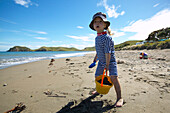 Young girl (2.5 y.) playing at the beach, wearing sun protection swim suit, in front of the tent, campground at Port Jackson on the northern tip of Coromandel Peninsula, North Island, New Zealand