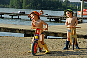 two boys with sunhat on juvenile bicycle, beach of lake Hartsee, Chiemgau, Upper Bavaria, Bavaria, Germany