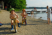 two boys with sunhat on juvenile bicycle, beach of lake Hartsee, Chiemgau, Upper Bavaria, Bavaria, Germany