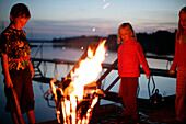 Three children standing around a fire near the jetty, evening, Lake Woerthsee, Upper Bavaria, Bavaria, Germany