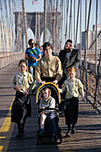 A severly-dressed Orthodox Jewish woman pushes a stroller across the Brooklyn Bridge in Manhattan, New York City. Note her husband in ethnic attire in right background and daughters in matching clothes.