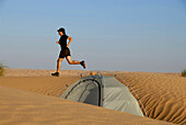 Woman jogging over dune, tent in foreground, Djebel Tembaine, Tunisia