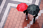 Two women with umbrellas walking in rain across brick paving stones. Westlake Center. Seattle. WA