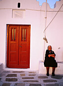 Greece, Cyclades Islands, Mykonos Senior lady looking having a rest in front of her door in a white washed street.