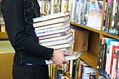 24 year old man standing, holding a pile of books, in the library