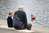 4 year old boy and his grandad, sailing a toy boat on a pond